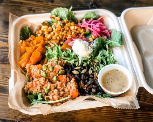 A colorful meal in a takeout container featuring greens, roasted vegetables, black beans, corn, pickled onions, and dressing.