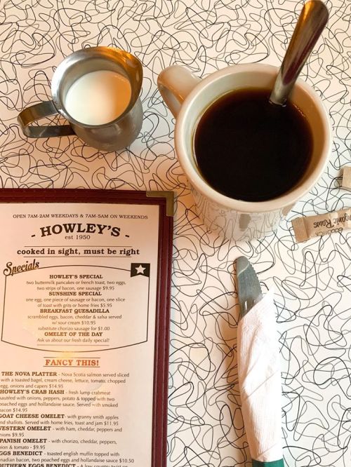 A table setting with a menu, coffee cup, cream pitcher, and utensils on a patterned tablecloth.