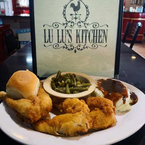 A plate of fried chicken, green beans, mashed potatoes with gravy, and a roll, with a menu sign for Lu Lu's Kitchen.