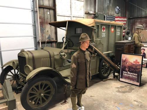 A vintage military vehicle displayed in a museum, alongside a mannequin in uniform and a historical poster.