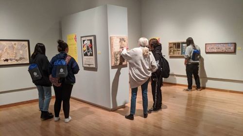 A group of visitors examines artwork in a gallery, with various framed pieces displayed on the walls.