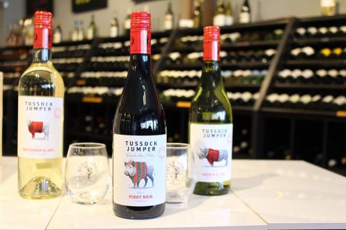 Two bottles of Tussock Jumper wine (Pinot Noir and Sauvignon Blanc) with glasses in front of a wine rack.