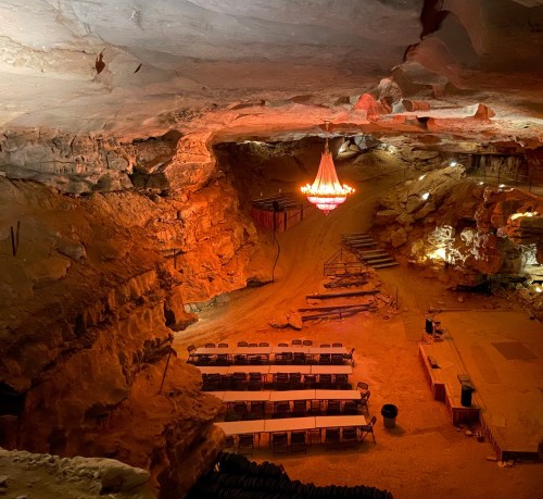 A cavernous space with a chandelier, featuring long tables and chairs set up for an event, illuminated by warm lighting.