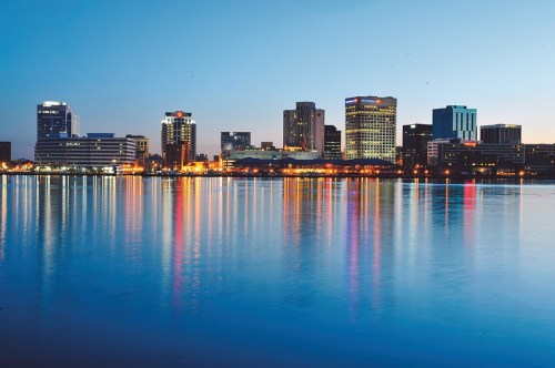City skyline at dusk, reflecting on calm water, with illuminated buildings and a serene atmosphere.