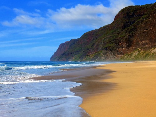 A serene beach with golden sand, gentle waves, and lush cliffs under a blue sky with scattered clouds.