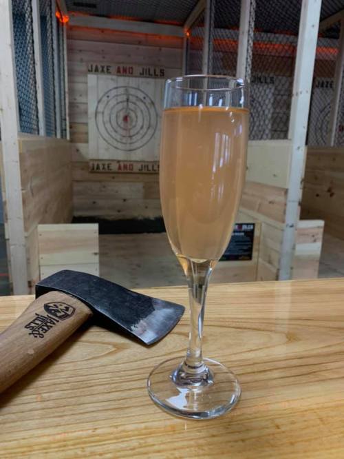 A glass of light pink drink on a wooden table, with an axe nearby and an axe-throwing target in the background.