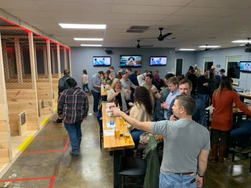 A lively indoor venue with people socializing, tables, and a wooden area for activities in the background.