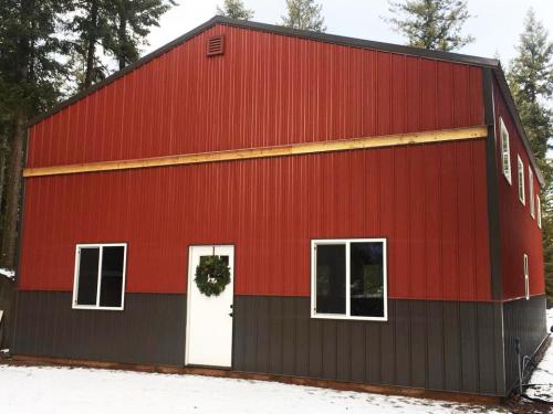 A red and gray metal building with a white door and windows, surrounded by snow and trees. A wreath hangs on the door.