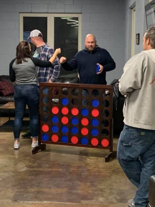 A group of people socializing around a large Connect Four game in a casual indoor setting.