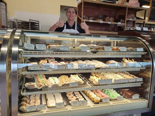 A smiling bakery employee gives a thumbs up in front of a display case filled with various pastries and desserts.