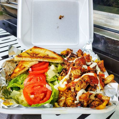 A takeout container filled with grilled chicken, lettuce, tomatoes, and pita bread, drizzled with sauce.