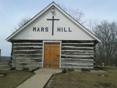 A log cabin church with "MARS HILL" written on the front, featuring a cross and a wooden door. Trees in the background.