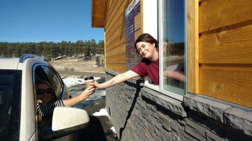 A barista hands a coffee to a customer through a drive-thru window on a sunny day. Snow is visible on the ground.