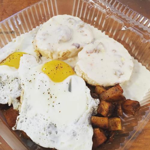 A takeout container with biscuits and gravy, two fried eggs, and seasoned potatoes.