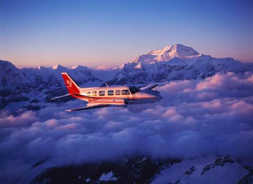 A small airplane flying above clouds with snow-capped mountains in the background during sunset.
