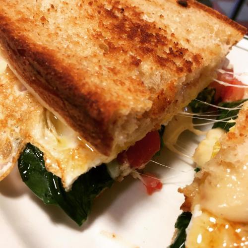 Close-up of a toasted sandwich with melted cheese, spinach, and tomato, showcasing gooey cheese stretching from the filling.