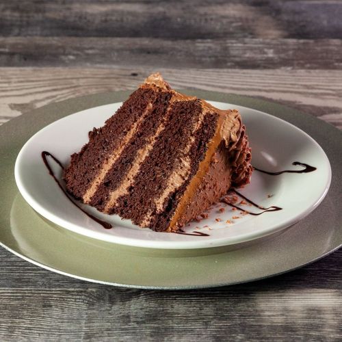 A slice of chocolate cake with layers of frosting on a white plate, drizzled with chocolate sauce.