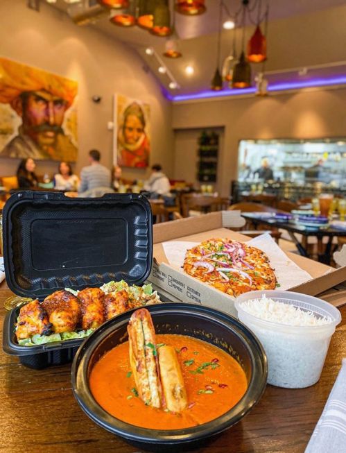 A takeout meal featuring chicken wings, curry, rice, and pizza, set in a cozy restaurant with colorful decor.