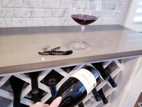 A hand holding a wine bottle near a glass of red wine and a corkscrew on a countertop with wine storage below.