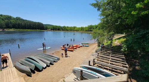 A sandy beach by a calm lake with kayaks and paddleboards, people enjoying water activities under a sunny sky.