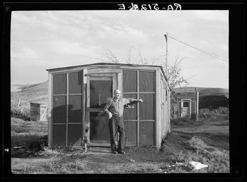 A man stands beside a small wooden shed in a rural landscape, with sparse vegetation and a cloudy sky in the background.