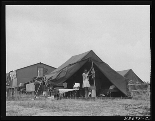 A person stands outside a large tent, with makeshift structures and a cloudy sky in the background.