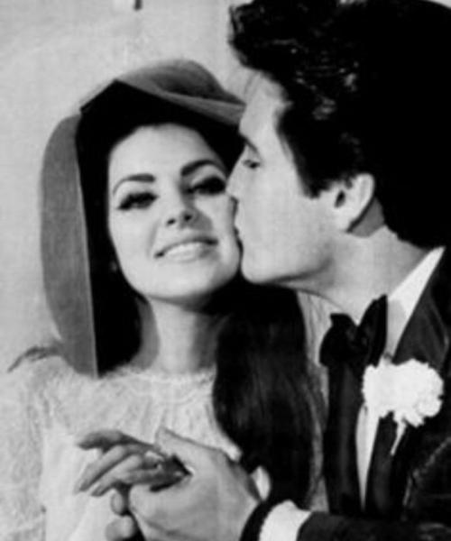 A black and white photo of a couple, the man kissing the woman's cheek as she smiles, both dressed elegantly.
