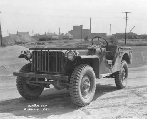 Black and white photo of a vintage military jeep parked on a dirt road, with industrial buildings in the background.