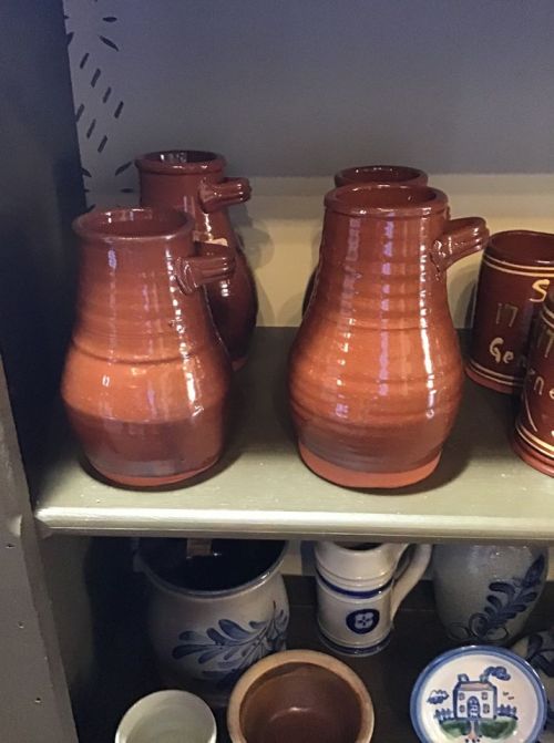 Three brown ceramic jugs with handles displayed on a shelf, alongside various other pottery items.