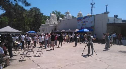 A crowd gathers outside a building with a colorful mural, surrounded by tents and trees on a sunny day.