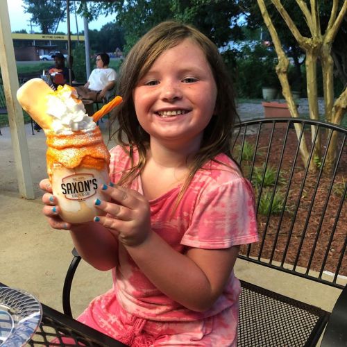 A smiling girl holds a colorful ice cream treat topped with whipped cream and a cookie, sitting outdoors.