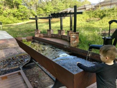 A child interacts with a water feature in a park, featuring a long, rust-colored basin and a pump. Greenery surrounds the area.