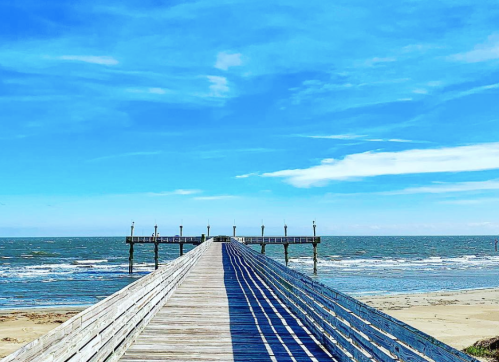 A wooden pier extends into the ocean under a bright blue sky with scattered clouds. Waves gently lap at the shore.