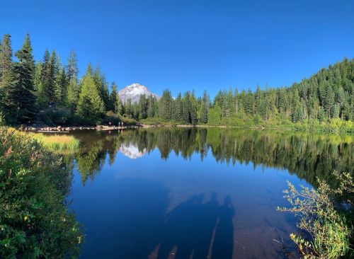 A serene lake surrounded by lush trees, reflecting a mountain and clear blue sky. Shadows of people are visible in the foreground.