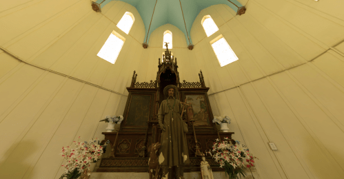 Interior of a small chapel featuring a statue of a saint, ornate altar, and floral decorations. Bright, airy atmosphere.