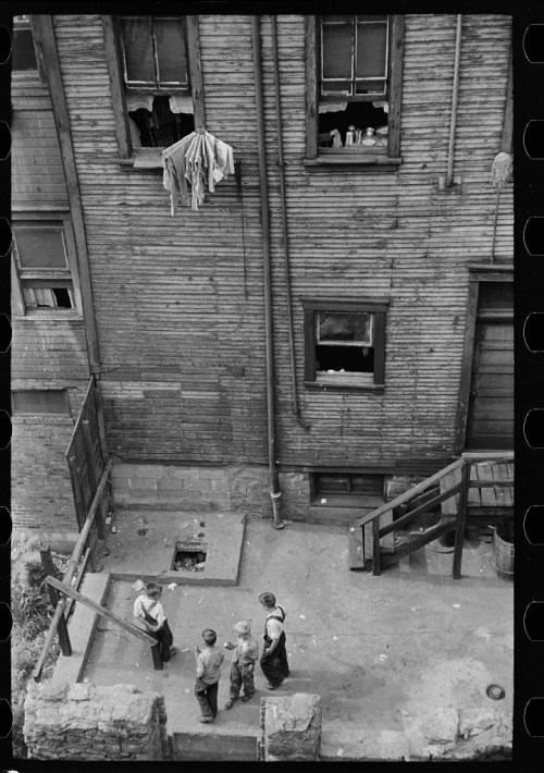 Children playing in a courtyard near a weathered wooden building, with laundry hanging from a window above.