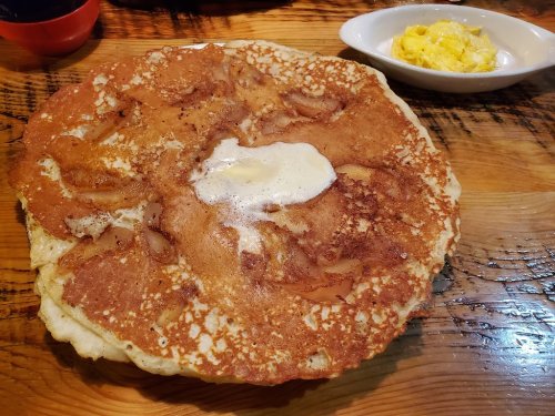 A large, golden-brown pancake topped with a pat of butter, served on a wooden table with a side of scrambled eggs.