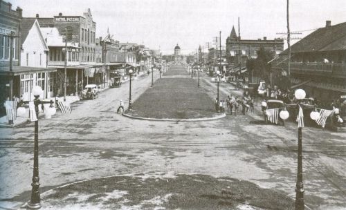 Historic black-and-white photo of a wide street lined with buildings, featuring people and decorative flags.