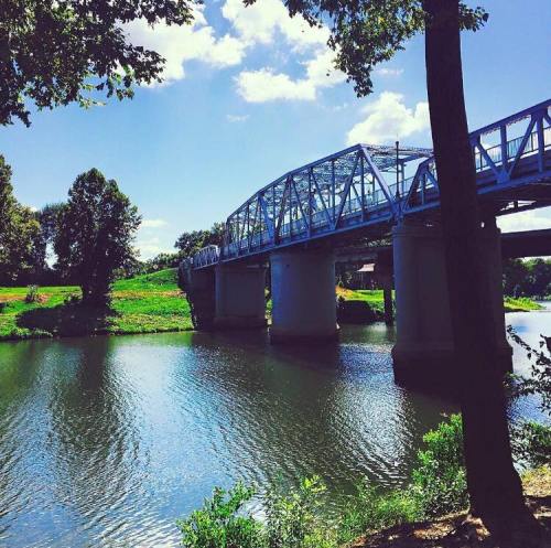 A blue metal bridge spans a calm river, surrounded by lush greenery and a clear blue sky with fluffy clouds.