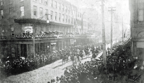 A historic street scene with a large crowd watching a parade, featuring horse riders and decorated buildings.