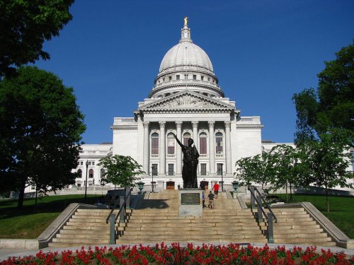 The Wisconsin State Capitol building with a large dome, flanked by trees and flowers, under a clear blue sky.