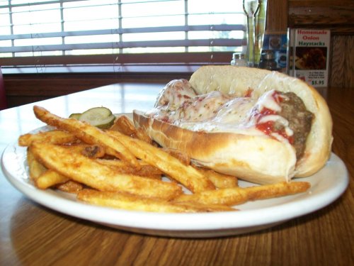 A plate with a meatball sub topped with cheese and ketchup, served with a side of golden fries and pickles.