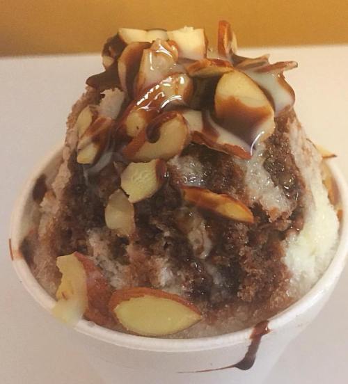 A cup of shaved ice topped with chocolate syrup, cake pieces, and sliced almonds.