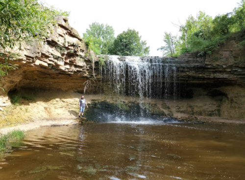 A person stands near a waterfall cascading over rocky cliffs into a calm pool surrounded by greenery.