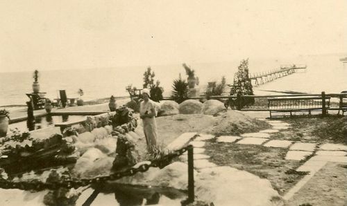 A vintage seaside scene featuring a man near a rocky landscape, with a pier extending into the water in the background.