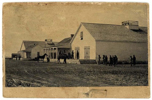Historic sepia-toned photo of a building with people and horses outside, set in a rural landscape.