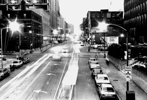 Black and white city street at dusk, with light trails from cars and vintage parked vehicles along the curb.