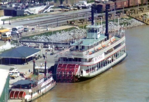 Two steamboats docked at a riverside, with a busy terminal and vehicles in the background.