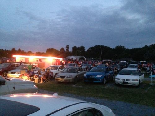 A crowded outdoor event at dusk, with many cars parked and people gathered near a brightly lit food stand.