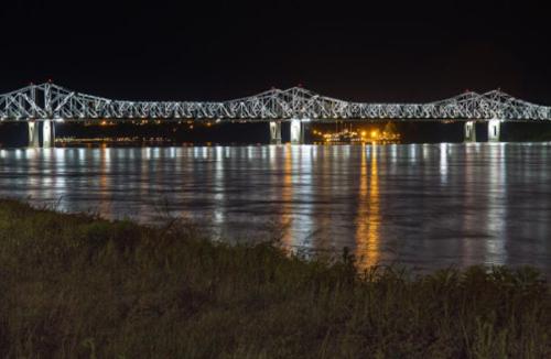 A brightly lit bridge spans a river at night, reflecting lights on the water's surface with a grassy foreground.
